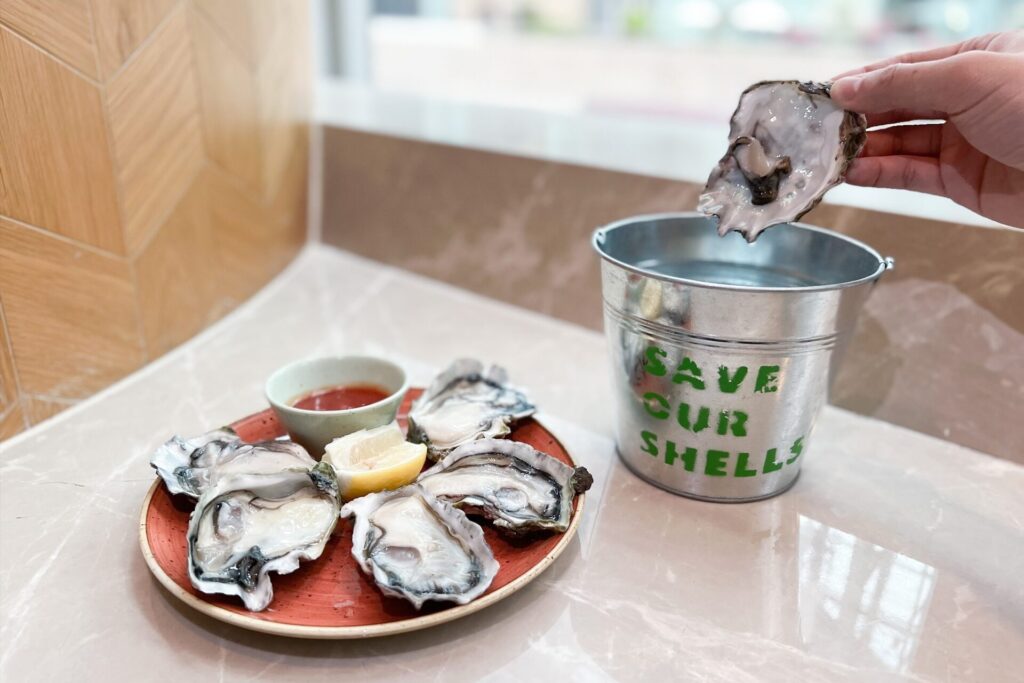 Oysters on a platter with a shell being placed into a silver bucket that says 'Save Our Shells'