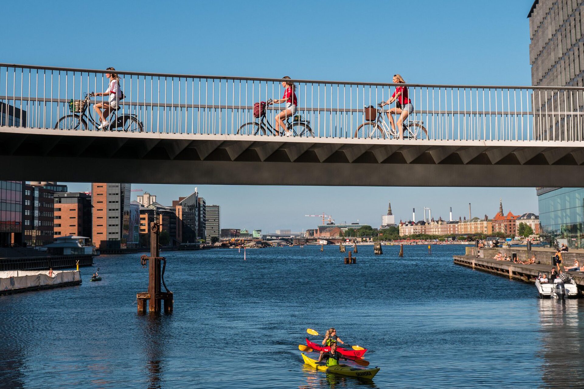 Three people riding over a bridge on bikes and two people canoeing under the bridge
