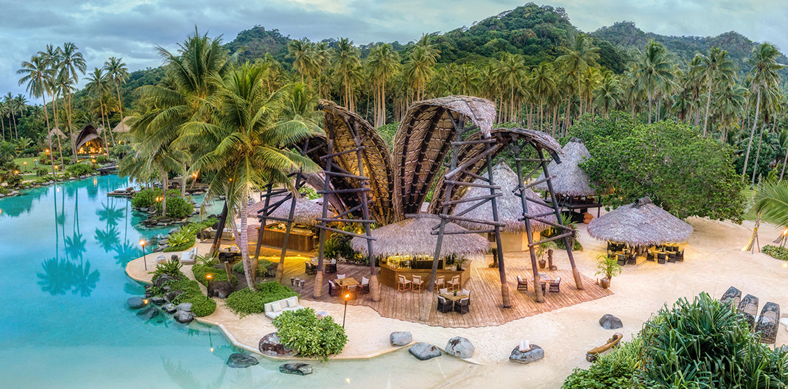 Aerial photo of COMO Laucala Island with multiple huts on a beachfront surrounded by trees.