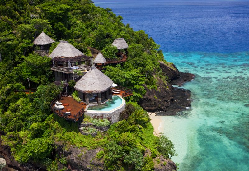 Aerial photo of COMO Laucala Island with multiple huts on a beachfront surrounded by trees.