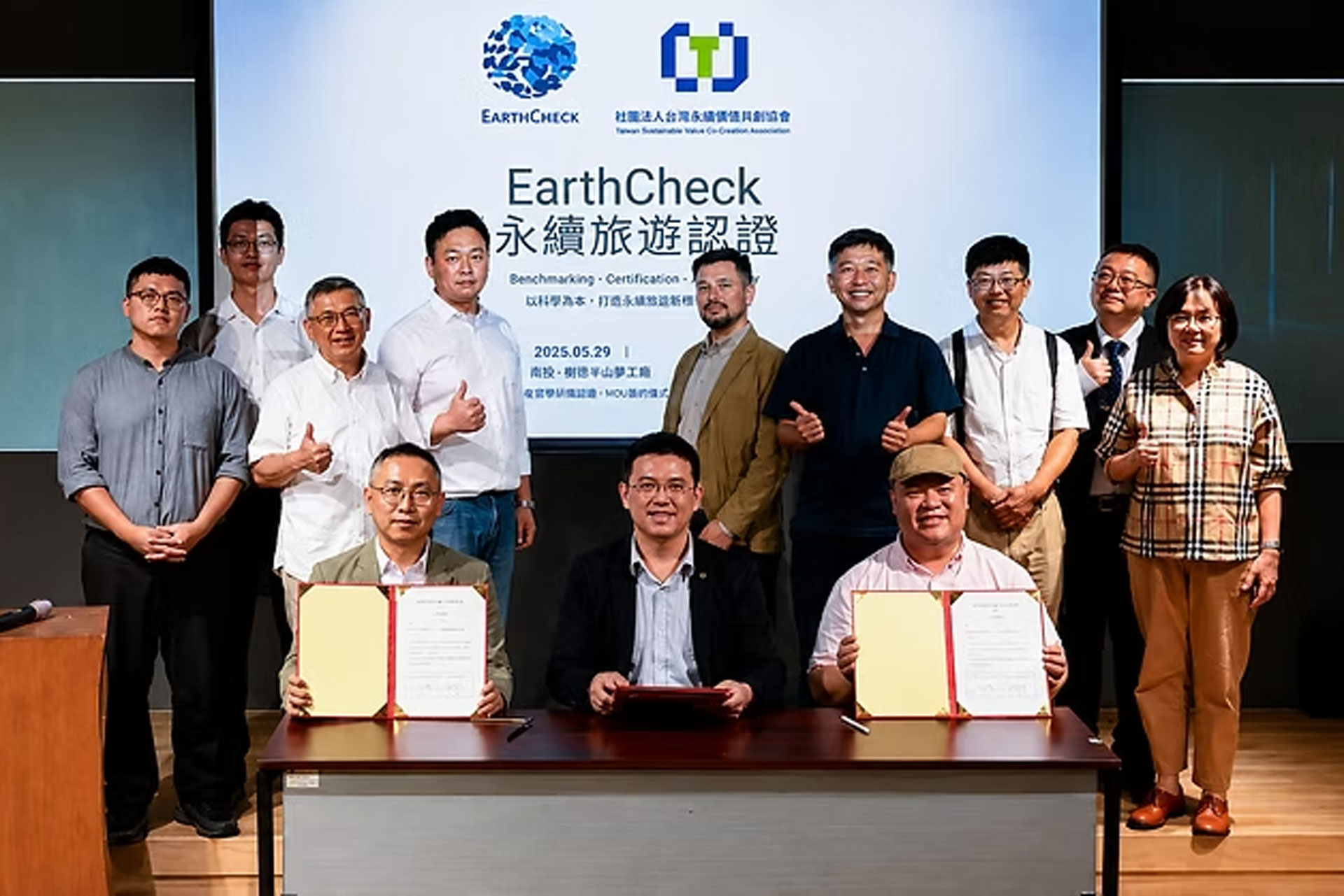Three people sitting at a table holding up certificates with a group of people standing behind them