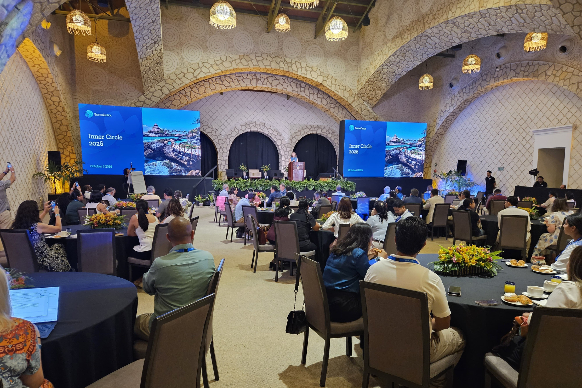 People sitting in groups around round tables while speaker presents at the front